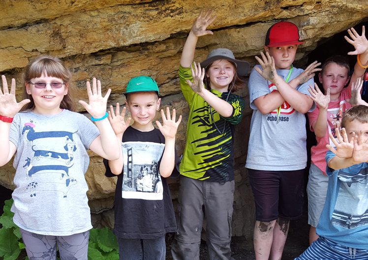 Group of children smiling and holding up hands outside of small cave