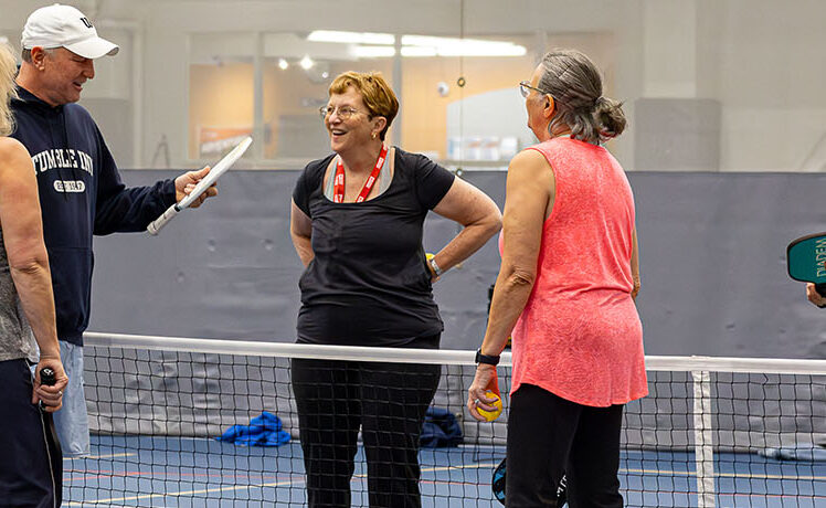 A group of four women and one man talking on a pickleball court.